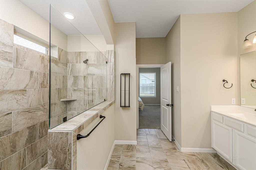 This bathroom features a spacious walk-in shower with elegant stone tile, a sleek glass partition, and a dual vanity with modern fixtures, all leading to a bright adjoining room.