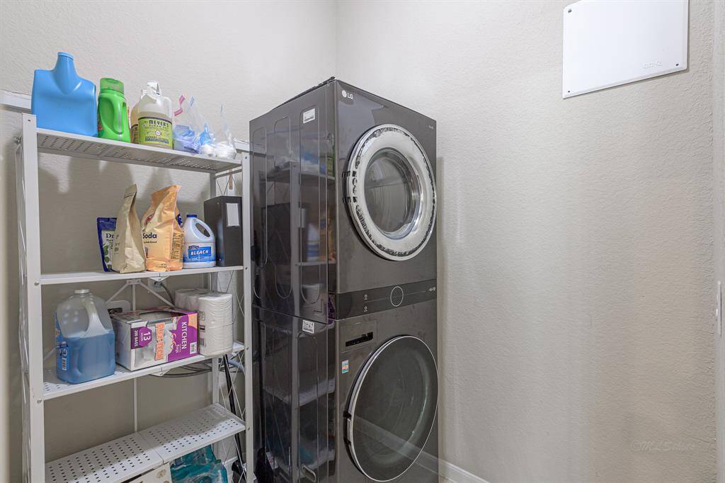 This photo shows a compact laundry room with a modern, stacked washer and dryer set. There's also a metal shelving unit stocked with various cleaning supplies, offering efficient storage space in a neutral-toned room.