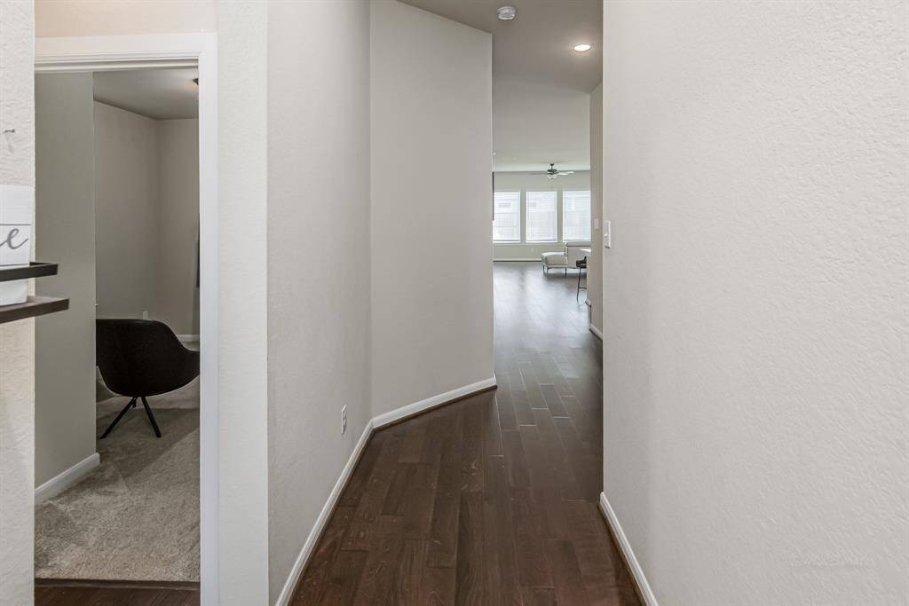 This photo shows a sleek hallway with dark wood flooring leading to a bright, open living area. There's a room on the left with carpet and a modern chair, suggesting a cozy study or office space.