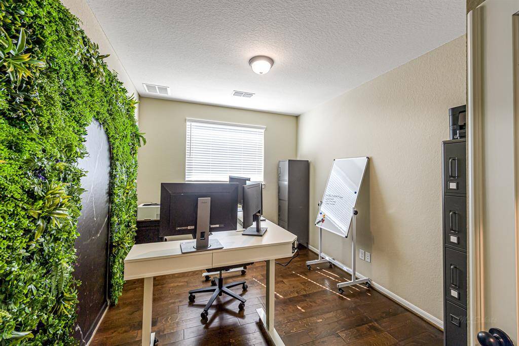 This home office features a modern setup with dual monitors on a sleek desk, a lively green plant wall, and ample natural light from a window. It includes storage cabinets and a whiteboard, creating a functional and inspiring workspace.
