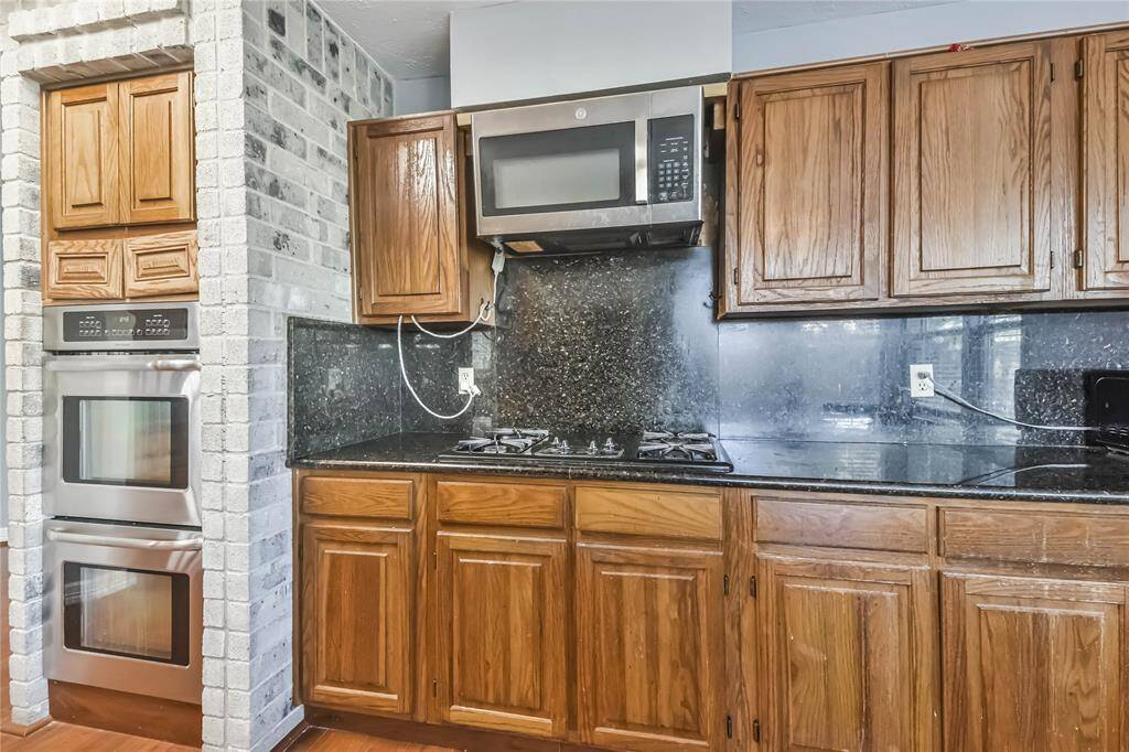 This kitchen features wooden cabinetry with granite countertops, a built-in microwave, a gas stovetop, and double stainless steel ovens. The brick accent wall adds a rustic touch.