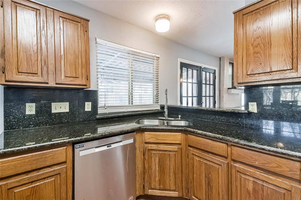 This kitchen features elegant dark granite countertops and natural wood cabinetry. It includes a stainless steel dishwasher and a double sink under a window, providing plenty of natural light. The space opens to a room with glass doors, enhancing the open feel.