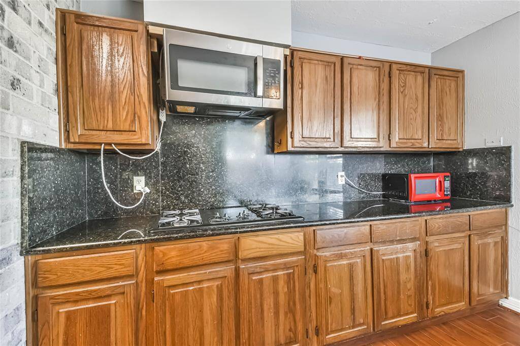 This kitchen features wooden cabinets, a sleek black granite countertop and backsplash, a built-in microwave, and a gas stove. There's a red microwave on the counter, and the room has a brick accent wall, adding character.