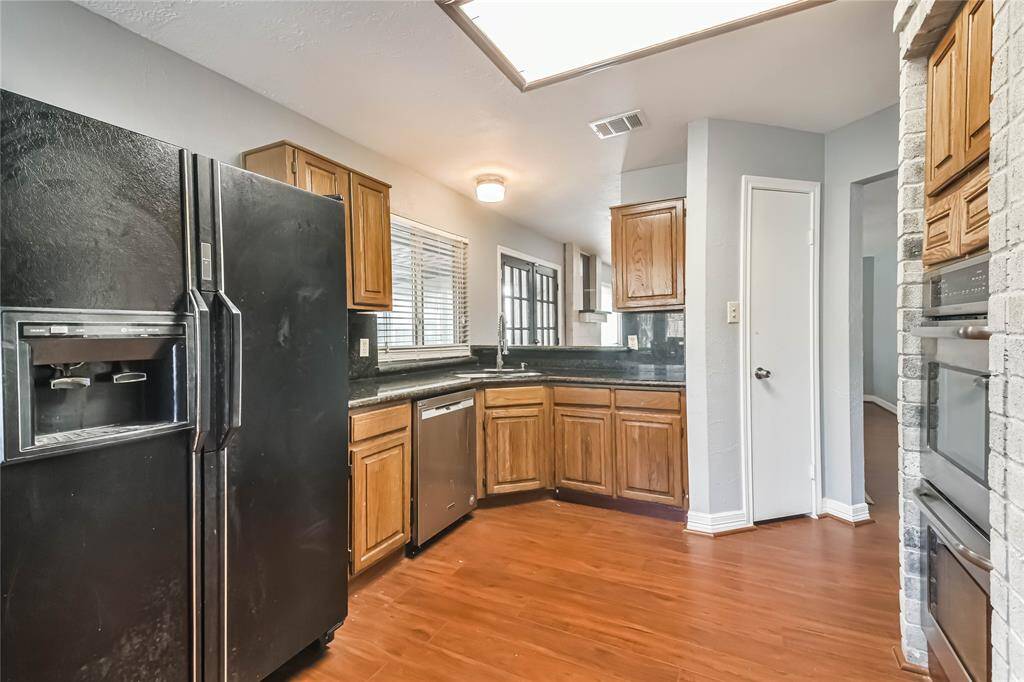 This kitchen features wooden cabinets, dark countertops, and black appliances, including a double-door refrigerator. It has a cozy layout with a window providing natural light and a brick accent wall housing built-in ovens.