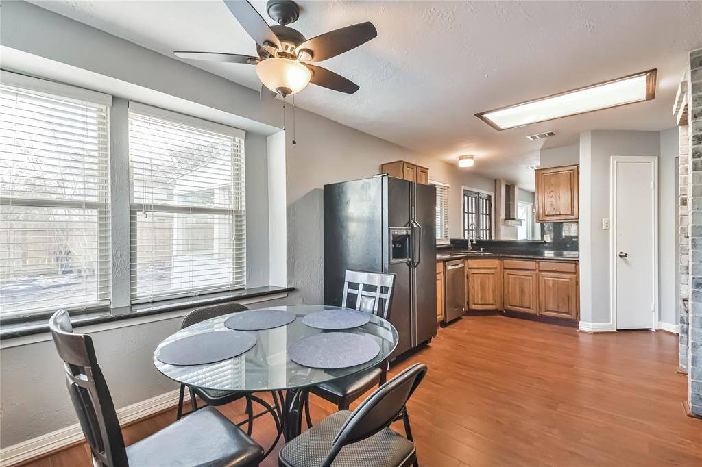 This bright kitchen features wooden cabinets, stainless steel appliances, and a cozy dining area with a glass table. Large windows and a ceiling fan enhance the space, offering a comfortable and inviting atmosphere.