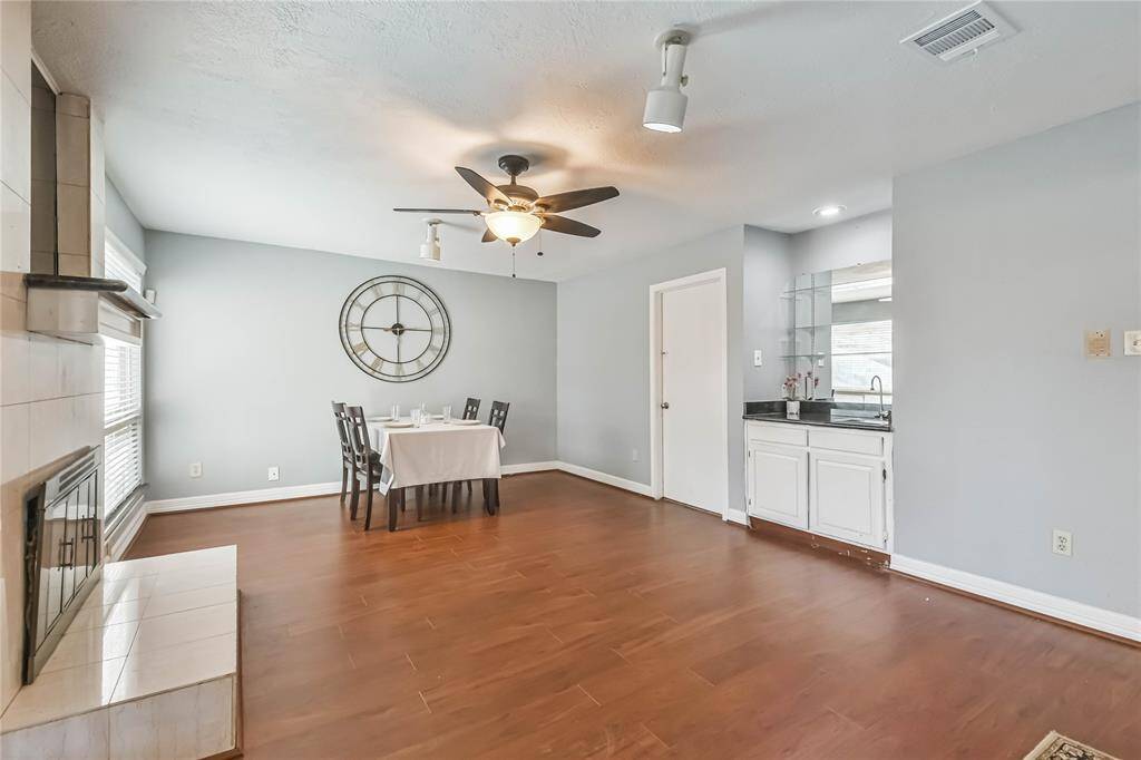 This inviting dining area features warm hardwood floors, a cozy fireplace, and a stylish ceiling fan. The space is well-lit with natural light from large windows. A small dining table is set up, and there's a convenient wet bar area with a sink and storage. The room is modern and functional, perfect for both entertaining and everyday dining.