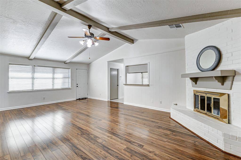 Welcome to your expansive living room, where a vaulted ceiling with exposed beams adds to the open, airy feel of the space. The clean white walls provide a bright, fresh backdrop, beautifully contrasting with the rich dark wood-look flooring, creating a perfect blend of elegance and warmth.