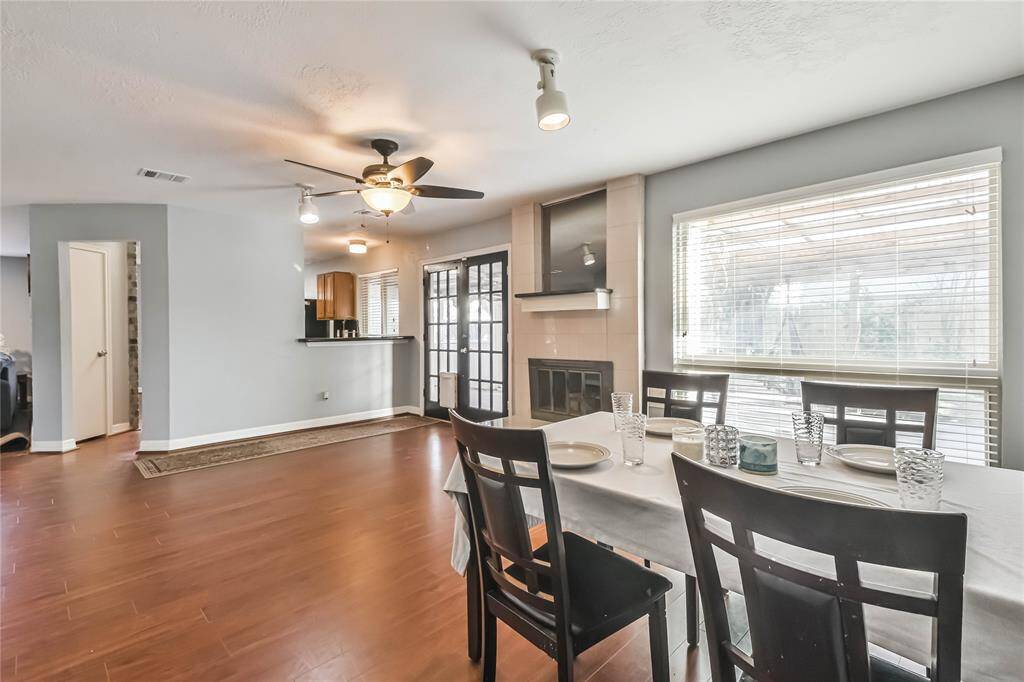 This photo showcases a bright dining area with wood flooring, a ceiling fan, and a large wall clock. There's a cozy fireplace on the side, and the room includes a small wet bar area with a sink. The space is open and inviting, perfect for entertaining.