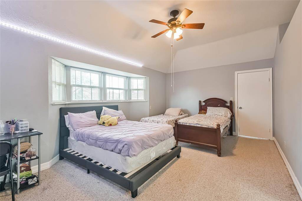 This spacious bedroom features neutral walls, a ceiling fan, and ample natural light from a large window. It accommodates two beds and has carpeted flooring, creating a cozy and comfortable environment.