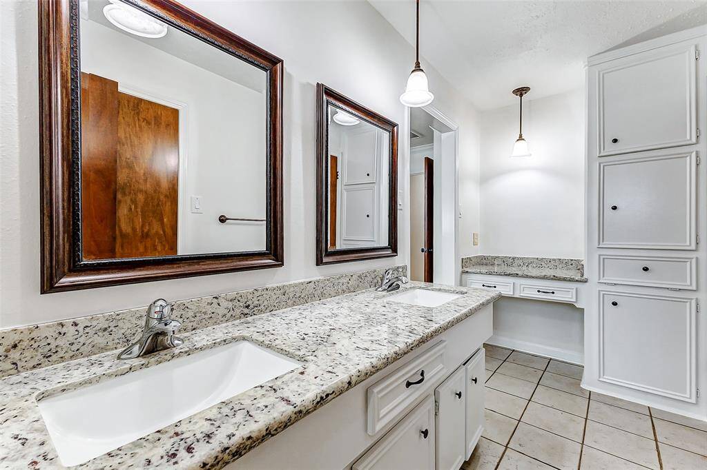 This ensuite bathroom boasts a double vanity with a sleek granite countertop and white undermount sinks, offering both style and functionality. Two dark-framed mirrors hang above, highlighted by pendant lights that add a touch of elegance. To the right, built-in cabinets offer plenty of storage, while the light-colored tile flooring adds a soft, neutral touch to the space.