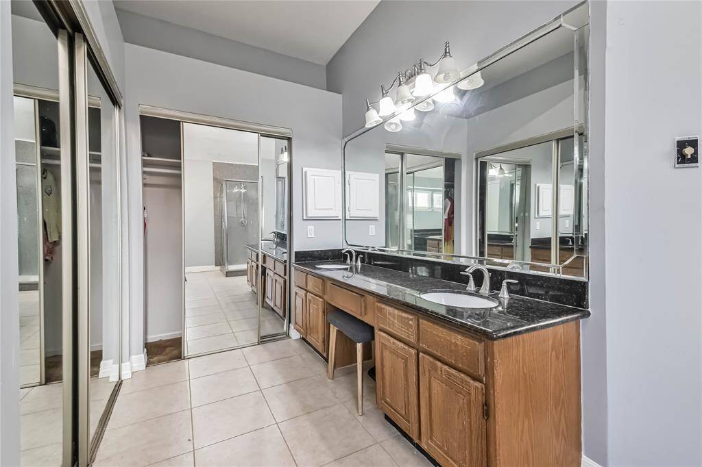 This spacious bathroom features a dual-sink vanity with granite countertops, large mirrors, stylish lighting, and tile flooring. It includes mirrored closet doors and a view into a modern shower area, offering a blend of functionality and elegance.