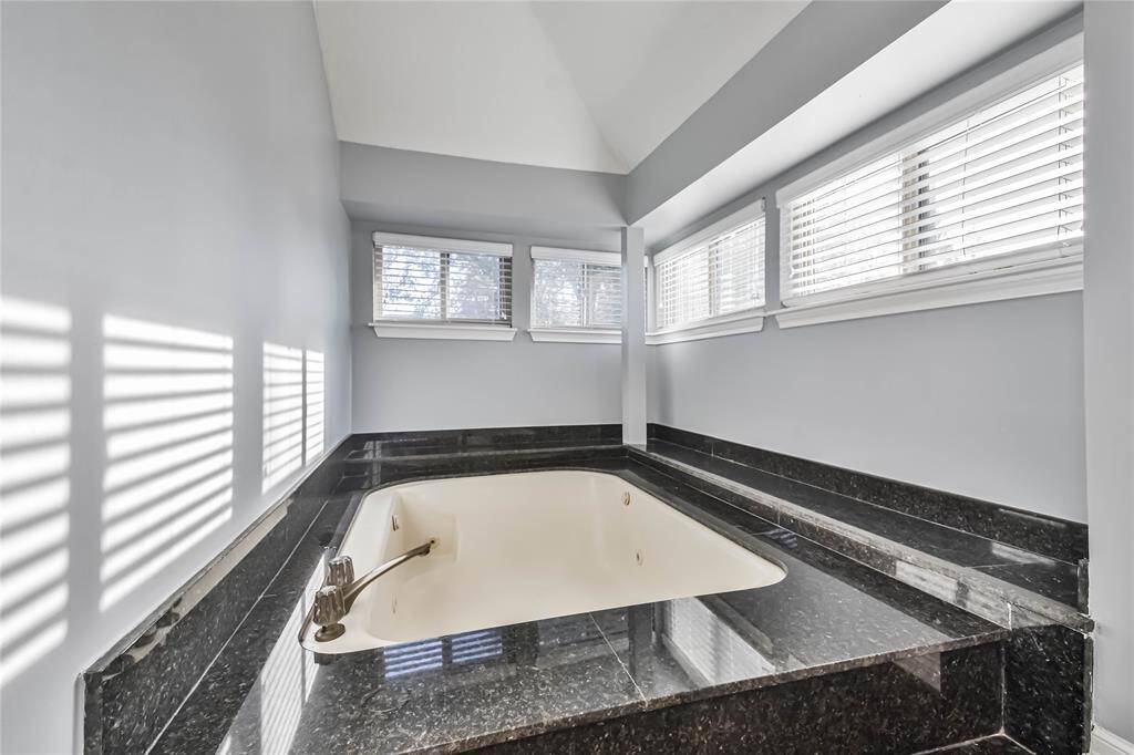 This photo showcases a luxurious bathroom featuring a sunken bathtub surrounded by elegant dark marble. The space is brightened by multiple windows with blinds, allowing natural light to fill the room, creating a serene and inviting atmosphere.
