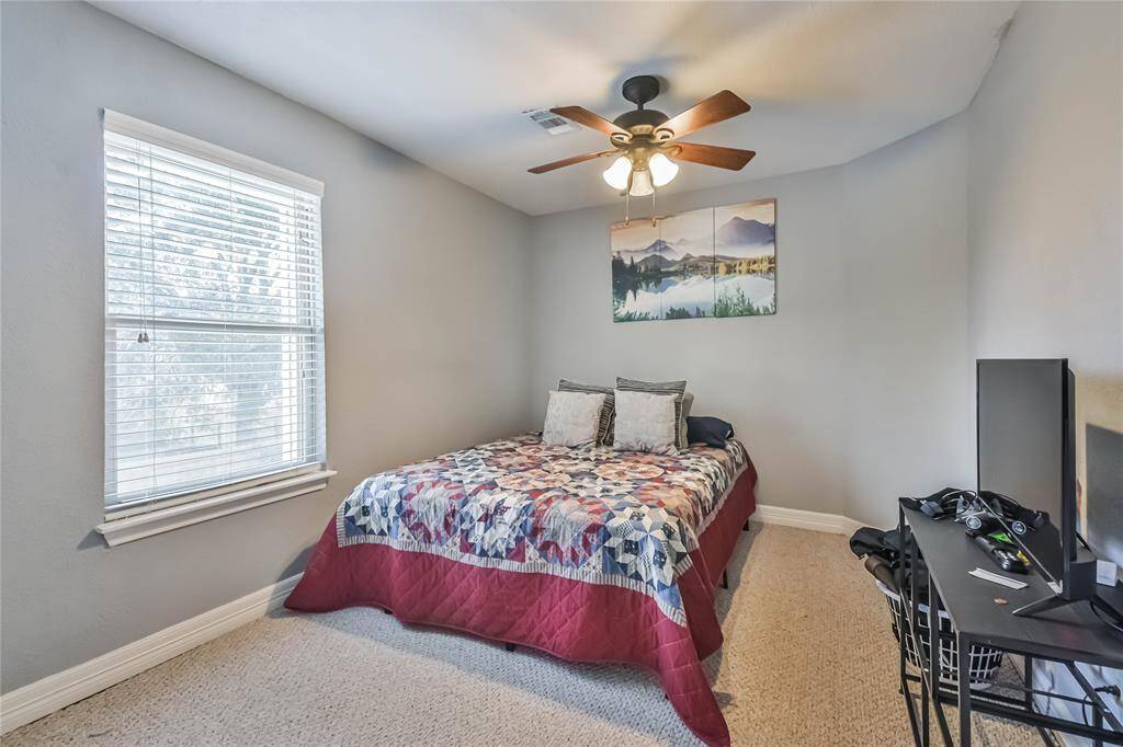 This cozy bedroom features soft gray walls, a ceiling fan, and ample natural light from a large window. It includes a comfortable bed with a colorful quilt and a small desk, perfect for a home office setup.