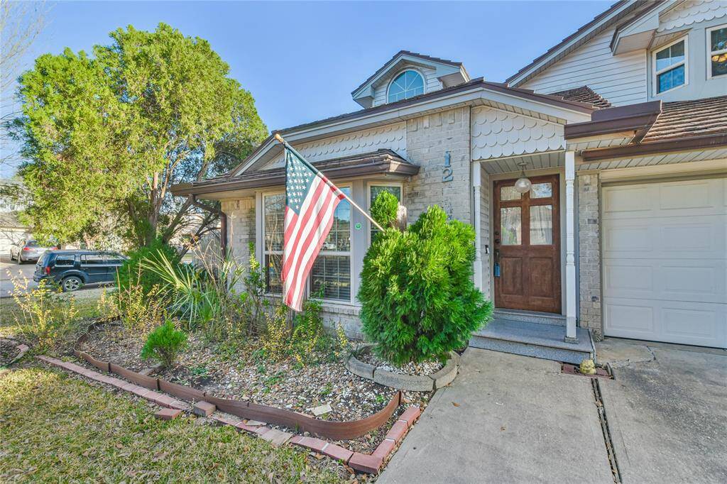 Charming brick home with a welcoming front porch, a well-maintained garden, and a one-car garage. The American flag and decorative details add character to the exterior.