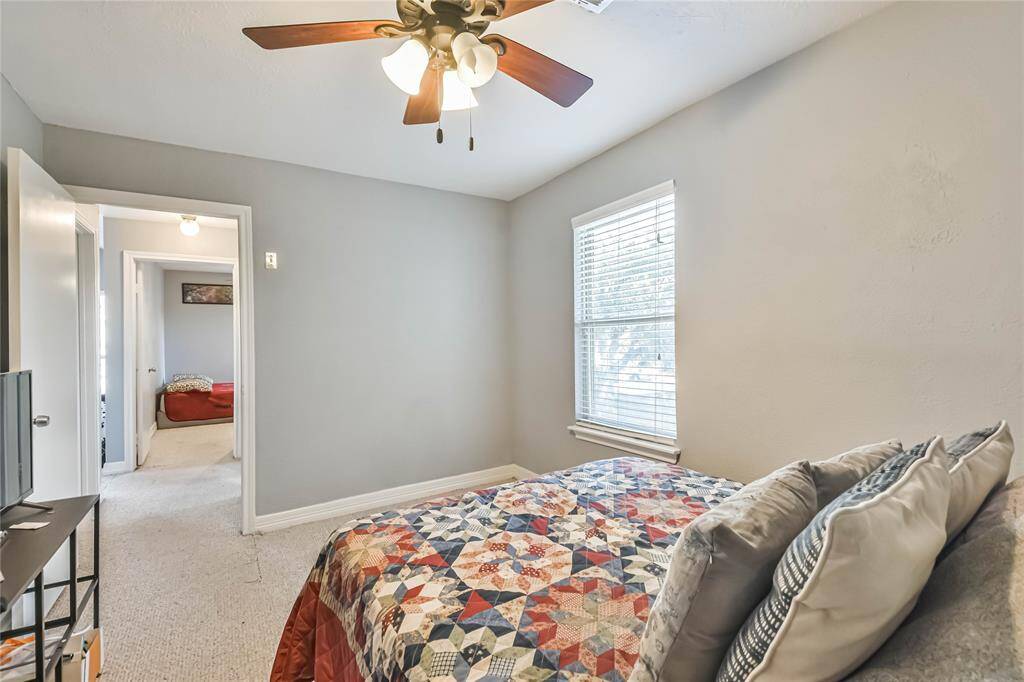 This is a cozy bedroom featuring a ceiling fan, a window with blinds, and neutral walls. The room is carpeted and includes a bed with a colorful quilt. A doorway leads to an adjoining room.