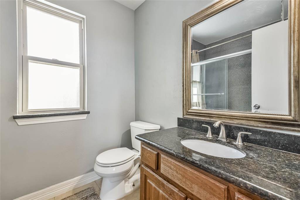 This is a modern bathroom featuring a sleek vanity with a granite countertop, a large framed mirror, and a window that provides natural light. It includes a toilet and a shower with a glass door, offering a clean and functional space.