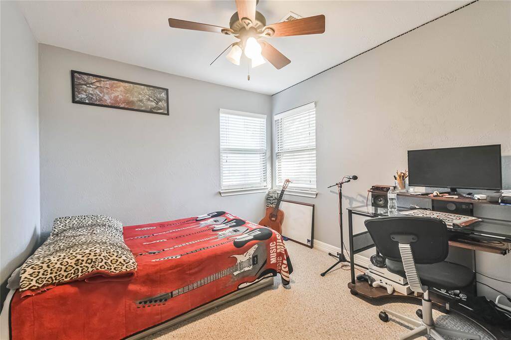 This cozy bedroom features neutral walls and carpet, with natural light streaming in through two windows. It includes a ceiling fan, bed, desk with a computer, and a guitar, creating a functional and inviting space.