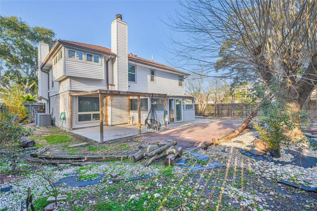 This photo showcases the backyard of a two-story home with a patio area and pergola. The exterior features light-colored siding and brick accents. The yard includes mature trees and a mix of grass and stone landscaping, creating a spacious outdoor area.