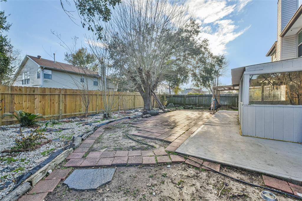 This backyard features a spacious patio area with paver stones, a mature tree providing shade, and a wooden privacy fence. There's a mix of landscaped and natural areas, offering potential for gardening or outdoor activities. A sunroom or enclosed porch is attached to the house, enhancing the outdoor living space.