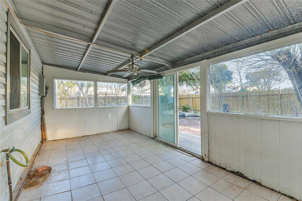 This photo shows a covered patio or sunroom with tiled flooring, large windows, and sliding glass doors leading to a fenced backyard. It features a ceiling fan and a metal roof, providing a versatile space for relaxation or entertaining.