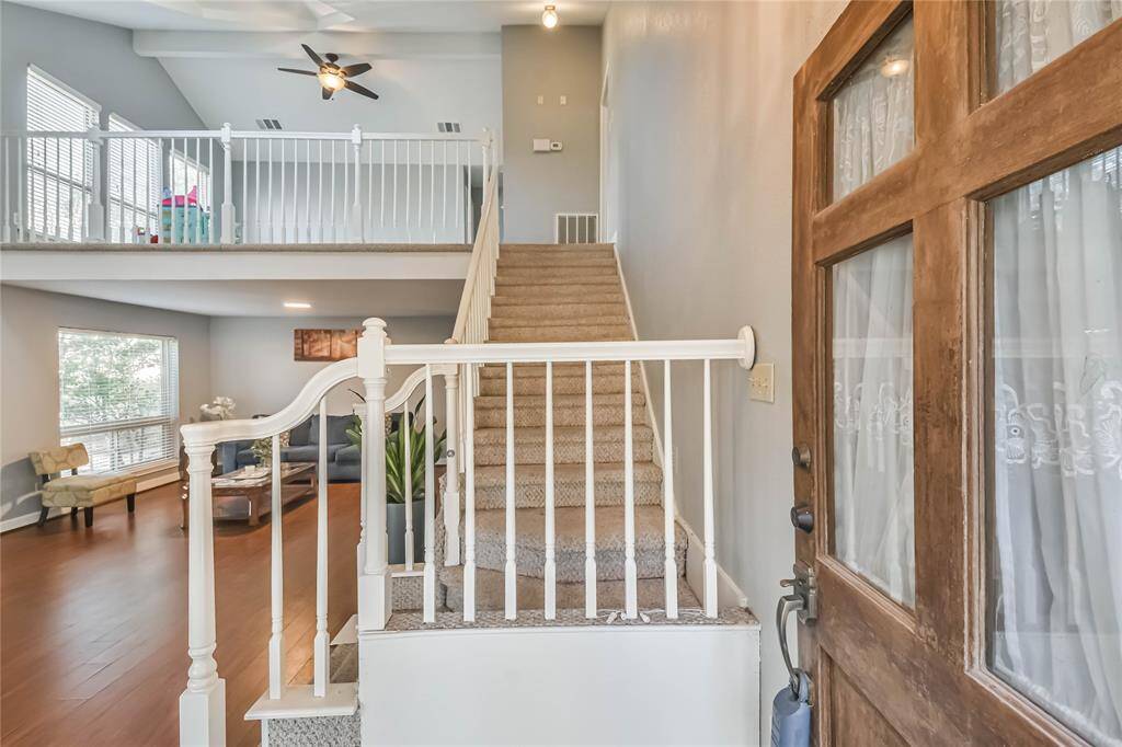 This inviting home features an open entryway with a wooden door and carpeted stairs leading to an upper level. The spacious living area boasts hardwood floors, large windows for natural light, and a ceiling fan, offering a cozy yet airy feel.