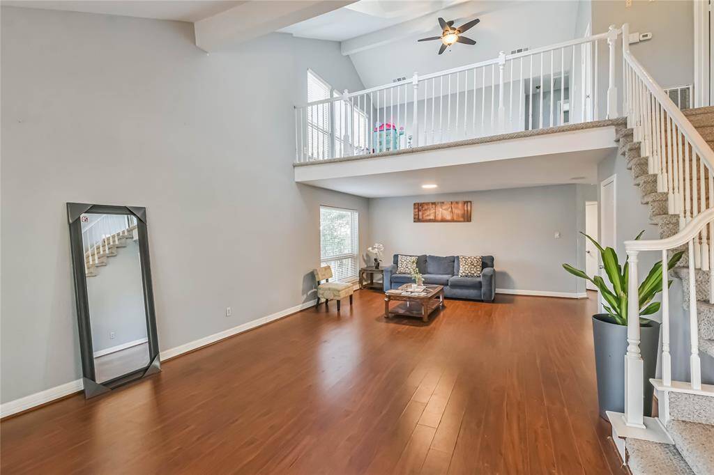 This spacious living room features a high ceiling with a large fan, an open loft area, and an elegant staircase. It has warm wood flooring, neutral walls, and ample natural light from a window by the sofa. Perfect for entertaining or relaxing.