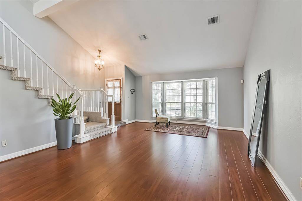 This spacious living room features warm wood flooring, a large bay window providing plenty of natural light, and a cozy seating area. The open staircase adds elegance, and a modern chandelier enhances the welcoming atmosphere.
