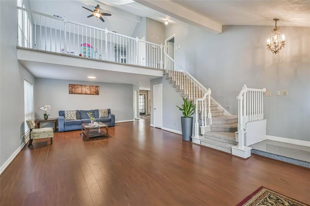 This spacious living area features high ceilings and abundant natural light. It includes a wooden floor, elegant staircase, and an open loft space. A modern chandelier and neutral decor add sophistication, making it an inviting space for relaxation and entertainment.