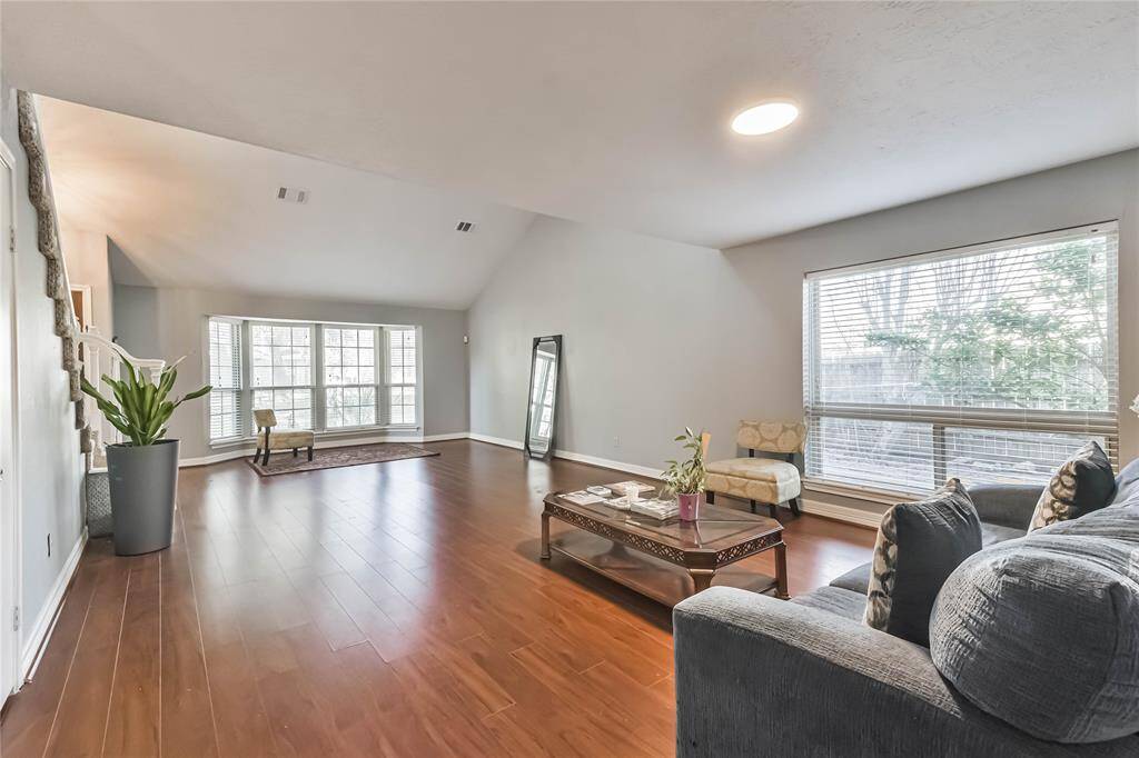 Spacious living room with large windows providing ample natural light, featuring hardwood floors and neutral walls. The area includes a cozy seating arrangement, a coffee table, and a staircase leading to the upper floor. Ideal for relaxation and entertaining.