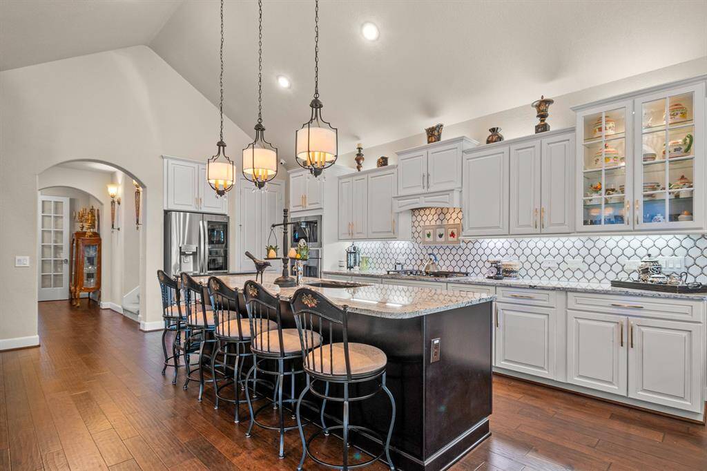 Discover this stunning Kitchen with an oversized with an extended island and additional seating, sleek stainless steel appliances, and elegant pendant lighting. Enjoy light cabinetry with glass displays, a decorative tile backsplash, and rich hardwood floors.