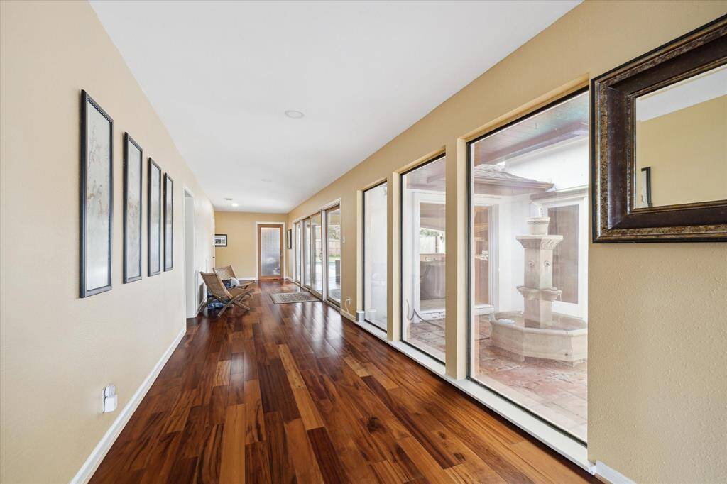 The bright, grand hallway with wooden flooring and a series of floor-to-ceiling windows allows views of the atrium with fountain, huge back covered patio with full kitchen, and pool.