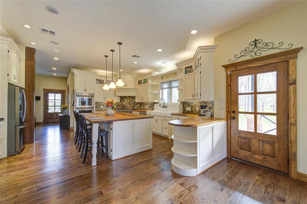 This kitchen features a warm, inviting atmosphere with rich wood floors and a large island with seating. It includes modern appliances, ample white cabinetry, and a charming farmhouse sink. Natural light streams through the glass-paneled door, enhancing the cozy, yet spacious feel.