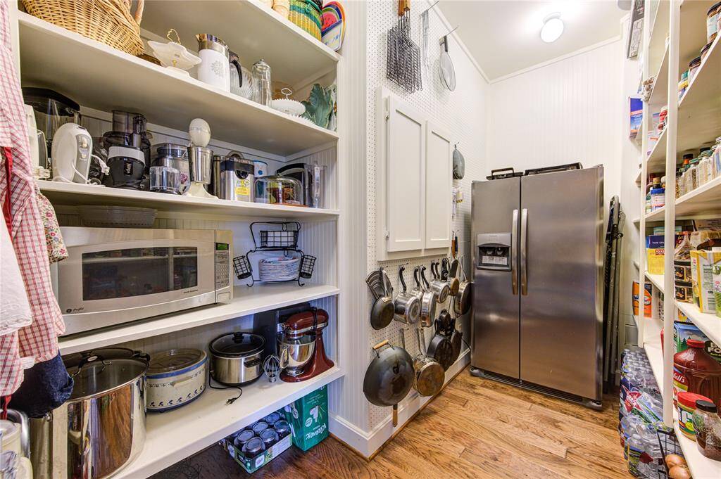 This is a dream of a pantry with ample shelving for kitchen appliances and groceries. It features a stainless steel refrigerator, wooden flooring, and wall hooks for pots and pans, maximizing storage and functionality.