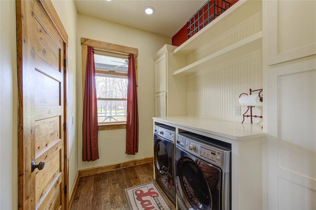 Spacious and functional laundry room with wooden flooring, housing a modern washer and dryer. It features ample storage space with built-in shelves and a window that brings in natural light.  A half bath is located right off this space for convenience!