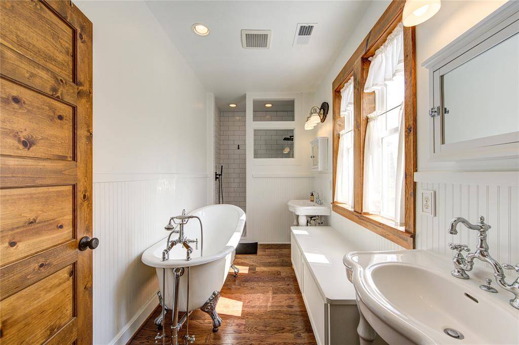 The primary bathroom features a vintage clawfoot tub, a separate shower with subway tiles, and his and hers classic pedestal sinks. It has warm wood accents, including a window seat, complemented by natural light from the large double windows. The space is finished with elegant wainscoting and hardwood floors and a mounted TV.