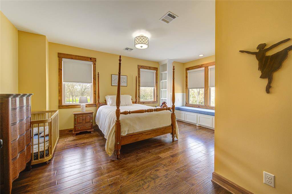 This cheerful secondary bedroom features warm yellow walls and rich hardwood floors. Large windows with wooden frames offer ample natural light and views of greenery. A built-in window seat with storage adds charm and functionality.