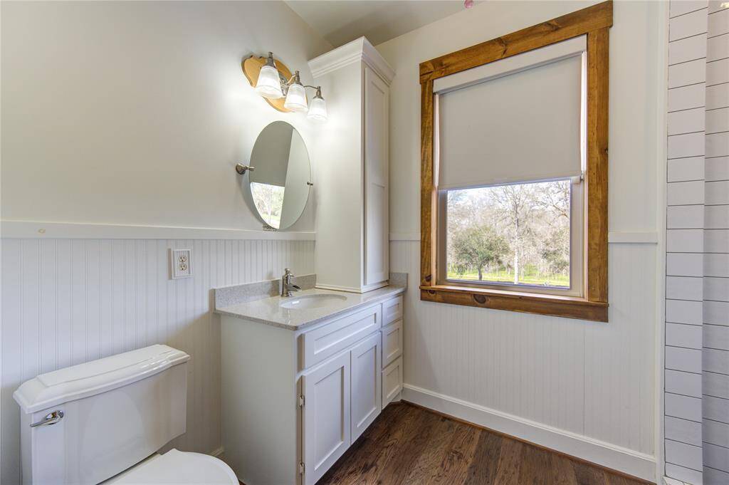 This en suite bathroom features a classic design with wainscoting and light walls. It includes a modern vanity with a quartz countertop, oval mirror, and polished chrome faucet. The room is well-lit with a sconce light fixture above the mirror. A large window with wooden trim offers natural light and a view of the outdoors, while a roller blind provides privacy. The flooring is rich hardwood, adding warmth to the space.