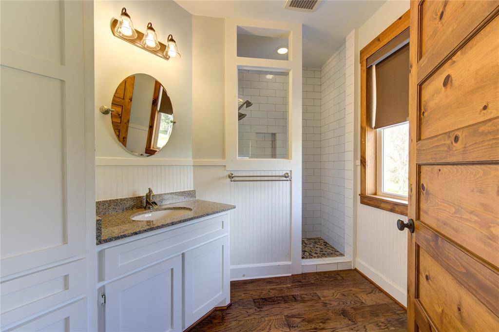 This en suite bathroom features a rustic design with wooden accents, a quartz countertop with a white vanity, and a glass-enclosed shower with subway tiles. It has a warm, cozy feel with ample natural light from the window.