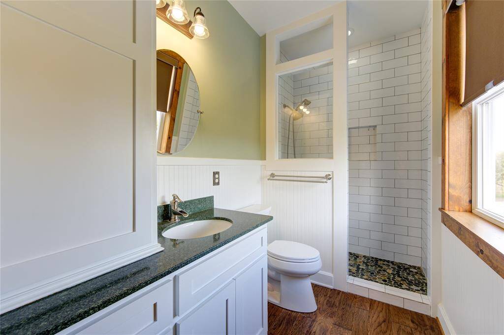 This en suite bathroom features a modern design with a sleek vanity, dark countertop, and round mirror. It includes a walk-in shower with white subway tiles and pebble flooring, complemented by wood flooring and natural light from the window.