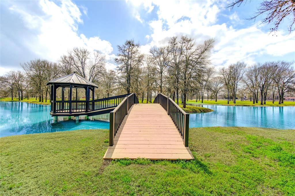 This serene property features a charming wooden bridge leading to a gazebo over a tranquil stocked pond, surrounded by lush green lawns and mature trees, offering a peaceful and scenic outdoor retreat.