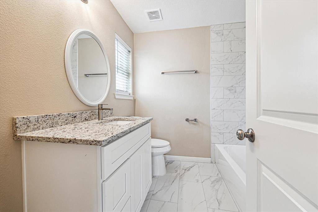 This Primary bathroom features a modern design with a marble-tiled shower, granite countertop, and white vanity. It is well-lit with a round mirror and has a neutral color palette for a clean, inviting look.