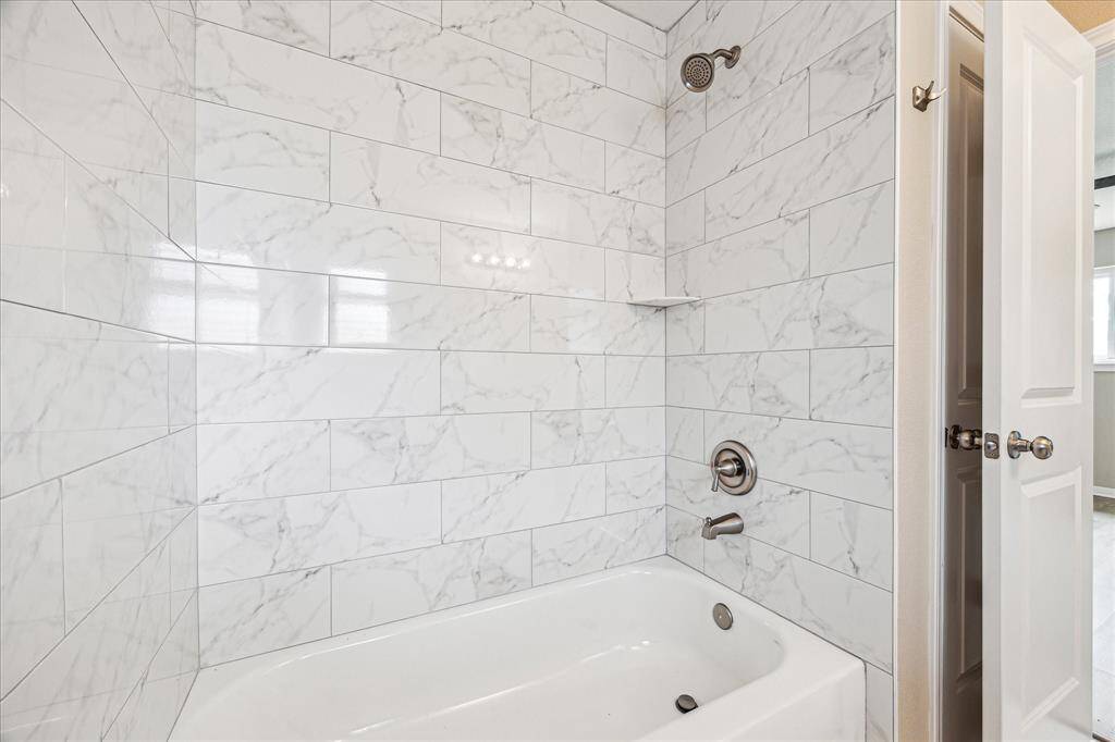 This bathroom features a sleek, modern design with white marble-style tile surrounding a bathtub and shower combination. It includes a chrome showerhead and faucet, complemented by a small built-in shelf for convenience.