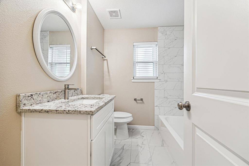 This hall bathroom features a modern design with a granite countertop, white vanity, round mirror, and tiled shower-bath combo. The neutral tones and natural light create a welcoming atmosphere. It is located across from the first secondary bed room.