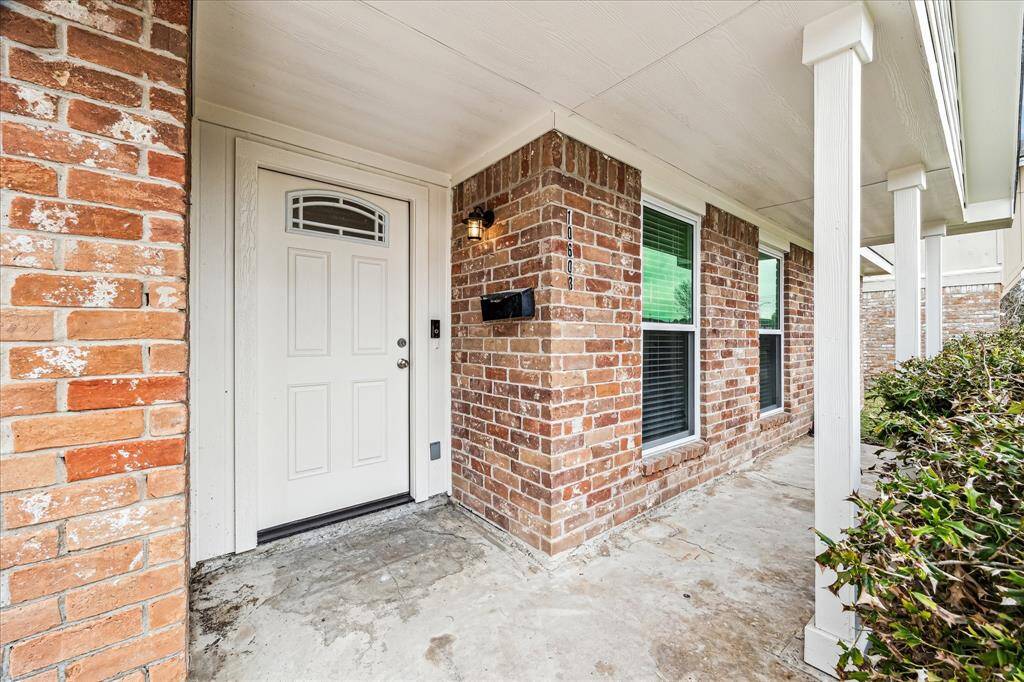 This photo shows a charming brick exterior with a covered porch area. The entrance features a white door with decorative glass and two windows providing natural light. There is greenery along the side, adding curb appeal to the welcoming front entry.