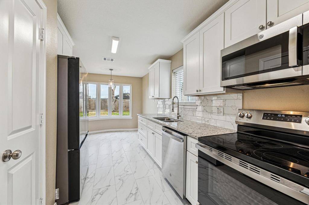 This kitchen features sleek white cabinetry, granite countertops, and modern stainless steel appliances. It has a bright, airy feel with a large bay window offering a view of the backyard. The space is enhanced by a marble-like tile floor and a stylish backsplash.