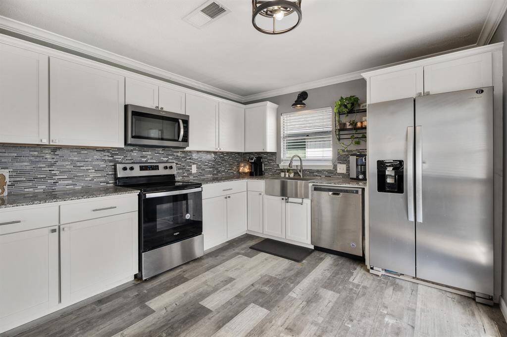 This modern kitchen features sleek stainless steel appliances, white cabinetry, a stainless steel farm sink and a stylish backsplash. The space is bright and inviting, with ample counter space and a double-door refrigerator for convenience.