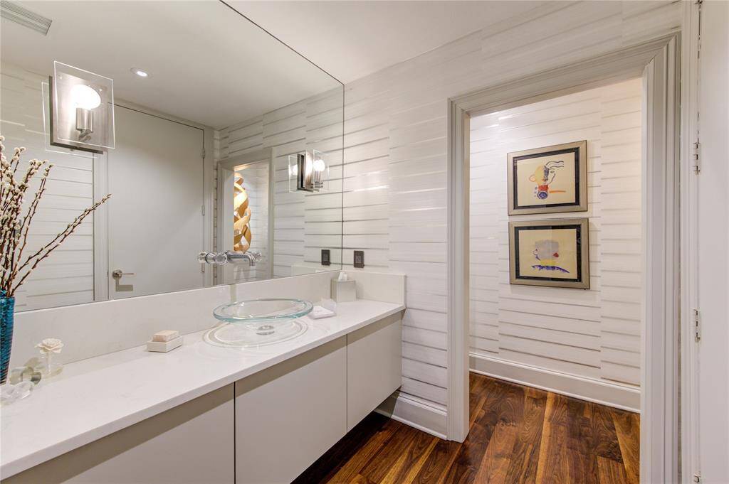 Downstairs powder bathroom features a modern design with white wall coverings, a sleek glass vessel sink, and contemporary light fixtures. The large mirror enhances the space, complemented by rich wood flooring. Closet not shown. This was originally a full bathroom.