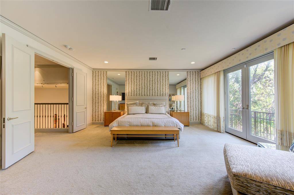 This luxurious bright bedroom has large windows and French doors providing abundant natural light and views of lush greenery. It features elegant wallpaper, plush carpeting, and a cozy bed setup. The room offers access to an small Juliet balcony through open double doors.