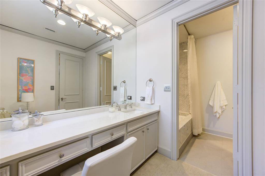 This hall bathroom features an updated design with a long vanity, a large mirror, and elegant lighting. It includes a marble-accented tub/shower and commode area.