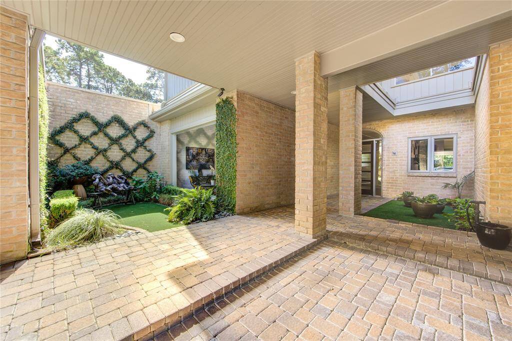 Another view of the charming courtyard with  brick pavers, lush greenery, and a trellis adorned with climbing plants. It features a covered walkway to the front door.